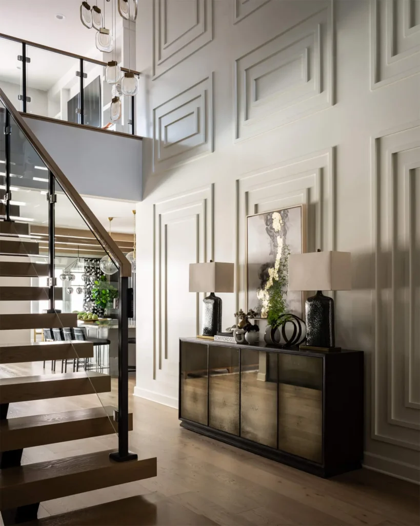 Modern staircase with open wooden treads and glass railing next to paneled white wall and console with lamps, designed by Grace Blu Interior Design.