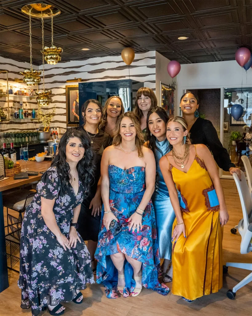 Group of eight women posing together in stylish dresses inside Grace Blu event lounge with zebra striped bar.