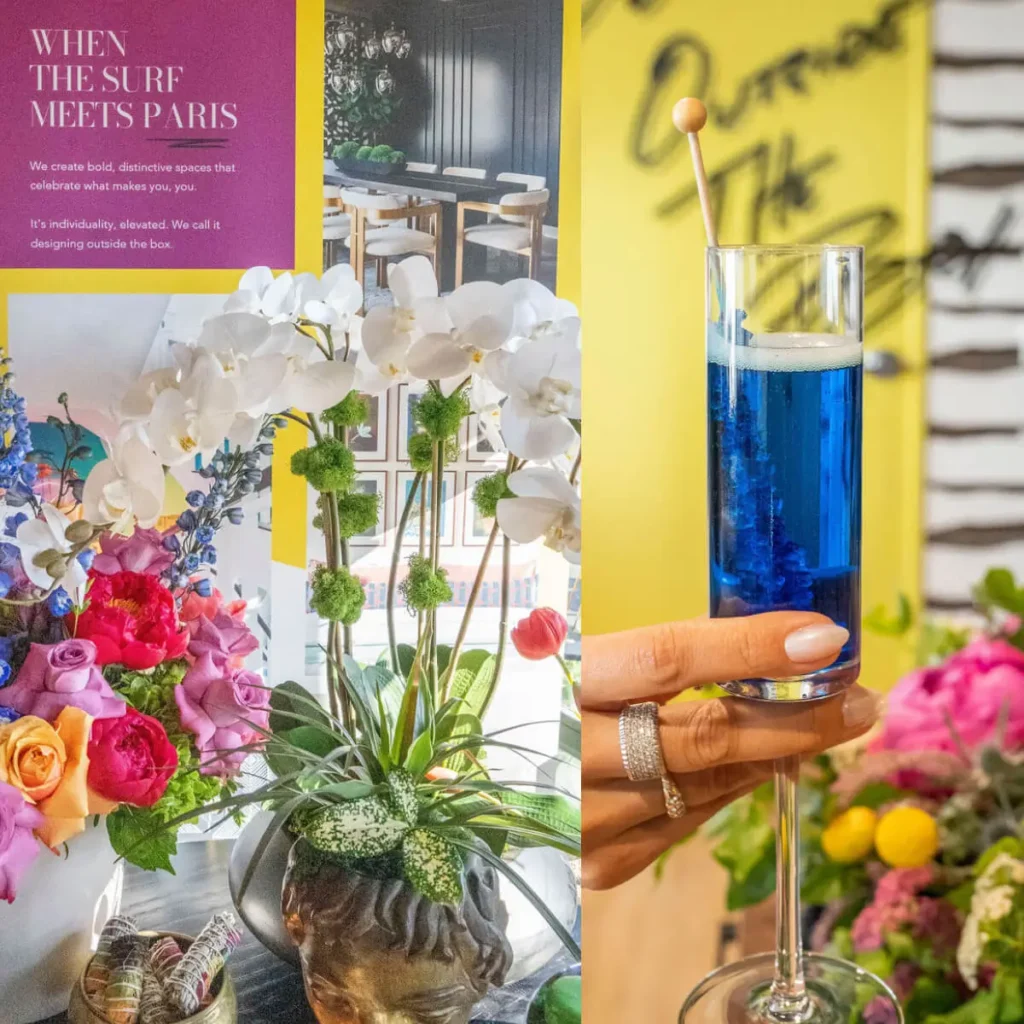 Hand holding a blue cocktail in front of vibrant floral arrangement and Grace Blu event poster reading 'When the Surf Meets Paris'.