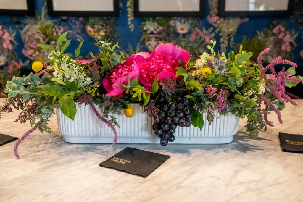 Lush floral arrangement with pink peonies, grapes and assorted greenery in white planter on marble table at Grace Blu event.