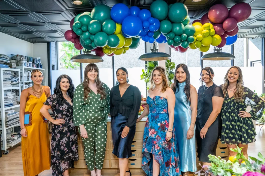 Group of eight women posing in colorful dresses under vibrant balloon arch inside Grace Blu studio during a celebration event.