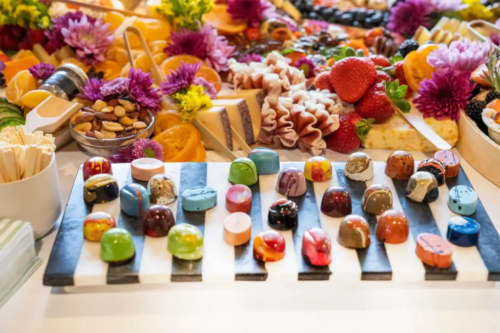 Assorted gourmet chocolates displayed on marble board with colorful charcuterie spread of meats, cheeses, fruits and flowers at Grace Blu event.