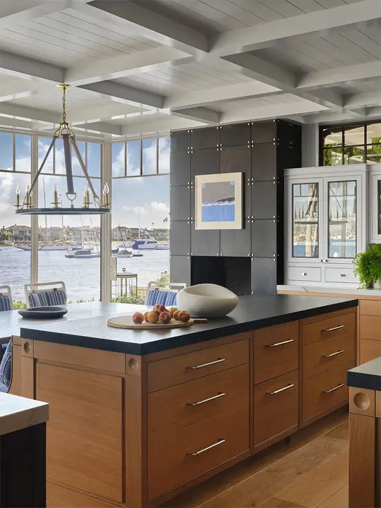 Kitchen from the Newport Nantucket project featuring a large wood island with black countertop, waterfront views through expansive windows, a coffered ceiling, and a black fireplace wall with framed art.