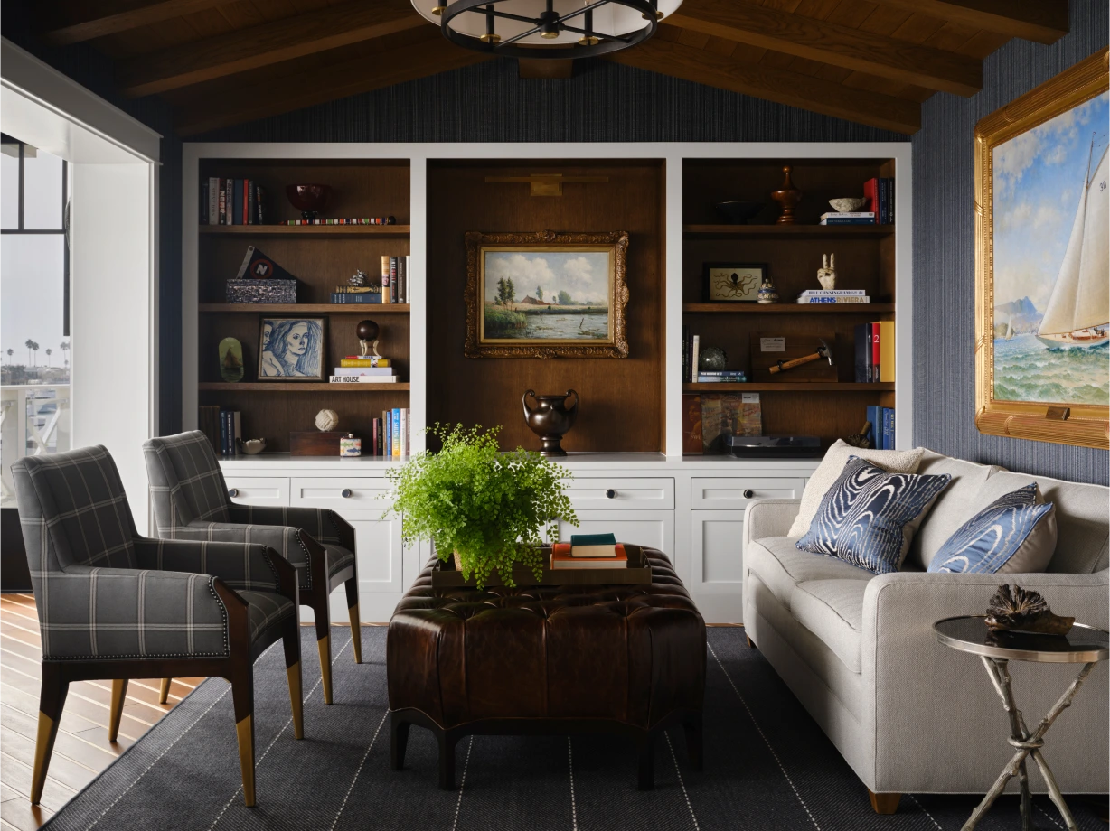 Cozy study with built-in bookshelves, plaid armchairs, leather ottoman, and nautical-themed artwork on the walls