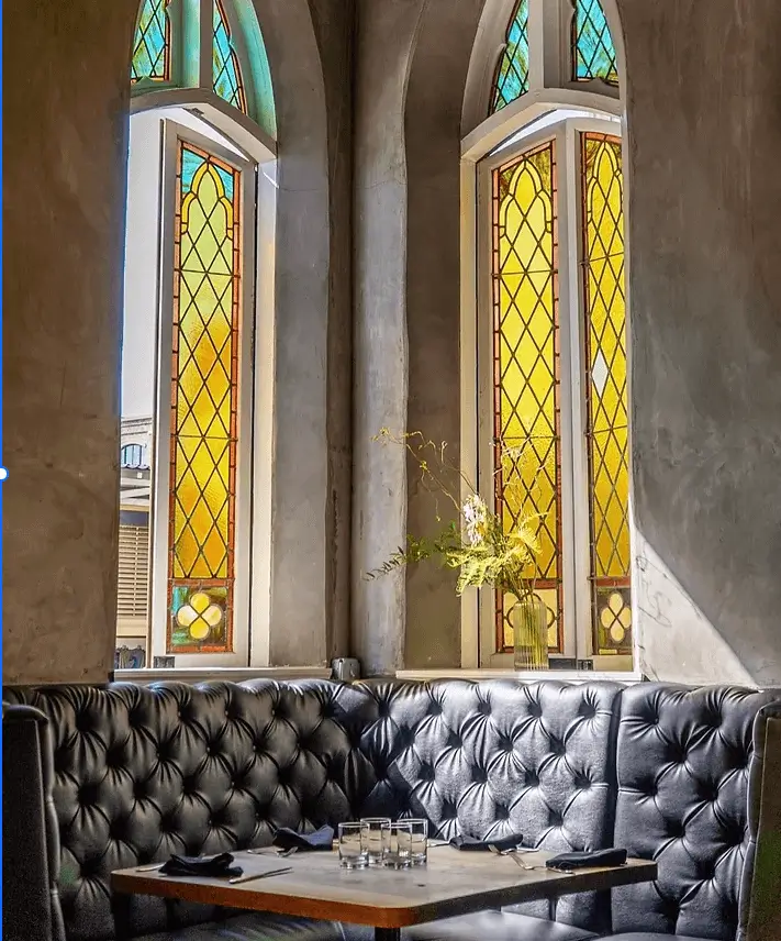 Cozy cafe booth with tufted black leather banquette beneath tall yellow stained-glass windows, capturing Grace Blu’s moody lounge inspiration.