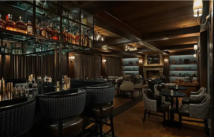 Moody lounge bar with dark wood paneling, mirrored shelves lined with liquor bottles, woven bar stools and ambient lighting, captured for Grace Blu.