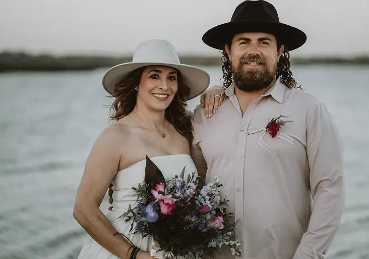 Couple wearing hats posing by the water, holding a bouquet of colorful flowers, representing Grace Blu lifestyle inspiration.