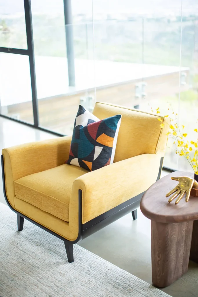 Yellow chair from Grace Blu’s Trailmaster Project styled with geometric pillow and a round wood side table with hand sculpture next to floor-to-ceiling windows.