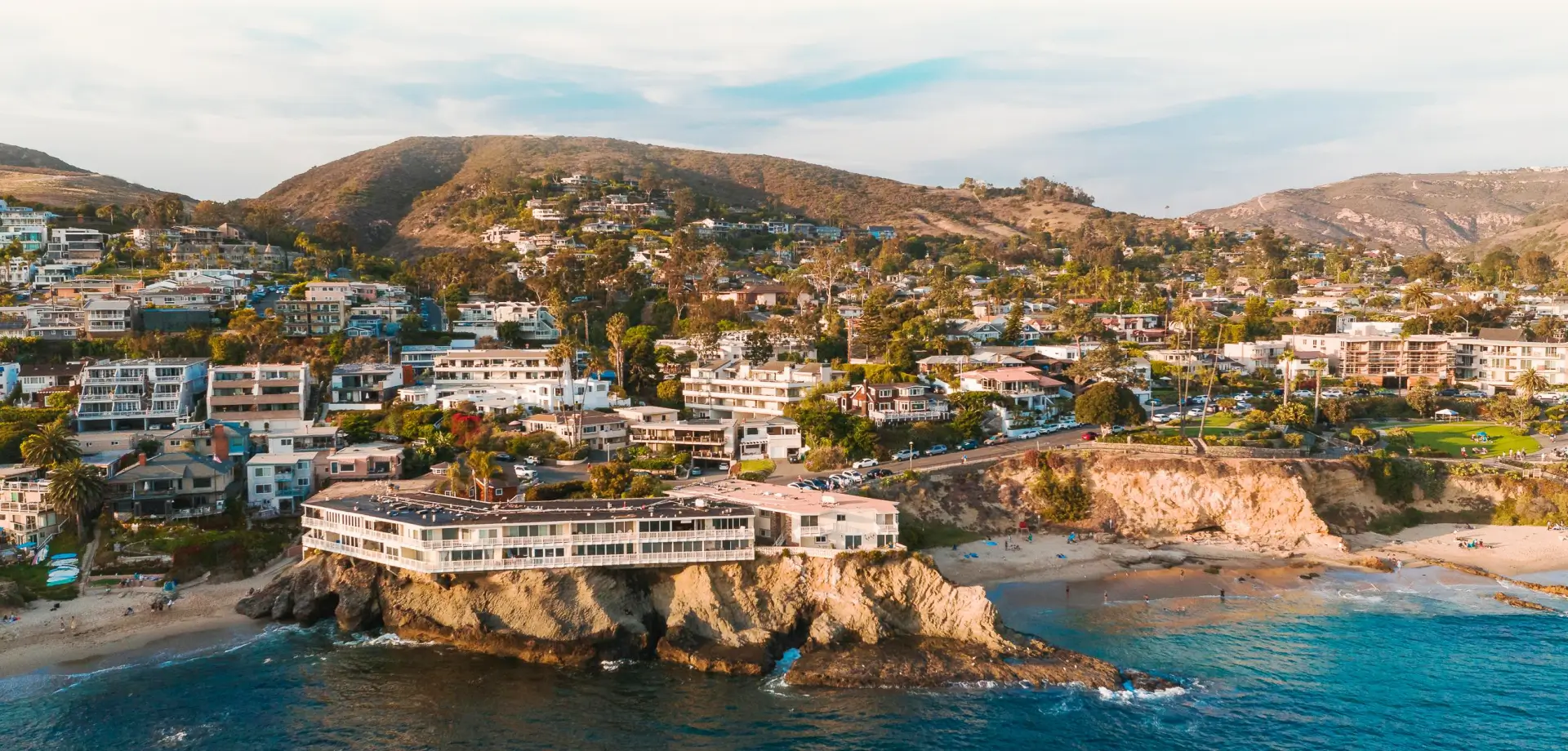 Aerial view of Laguna Beach coastline with rocky cliffs, beach houses, and hills at sunset for Grace Blu Interior Design