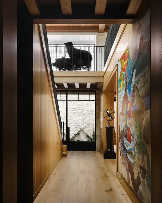 Hallway from the Newport Nantucket project featuring warm wood paneling, an eclectic colorful mural, natural stone wall at the far end, and a grand piano visible on the upper level.