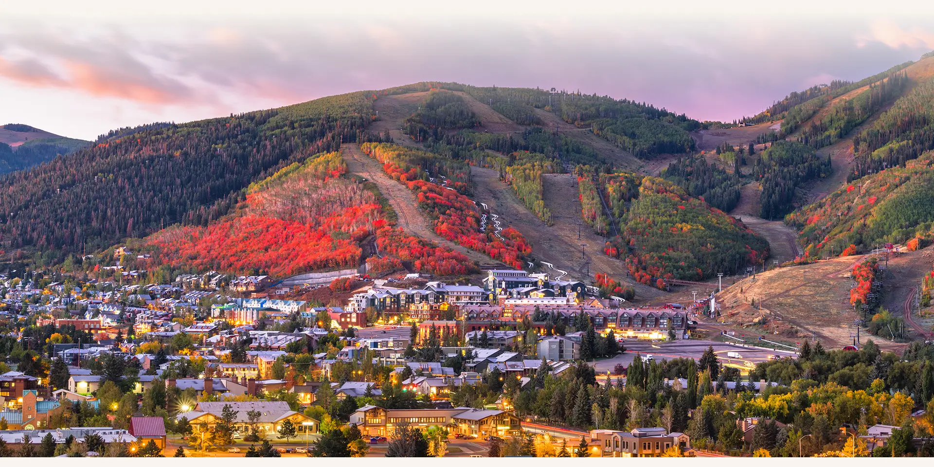Park City sunset with glowing city lights, ski lifts, and fall-colored trees.