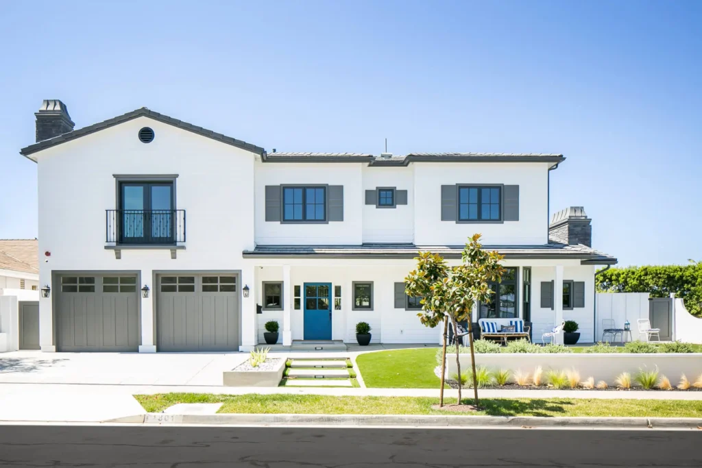 A bright, two-story white house with a modern farmhouse architectural style. The house features a gray tiled roof, gray shutters on the windows, and two gray garage doors.