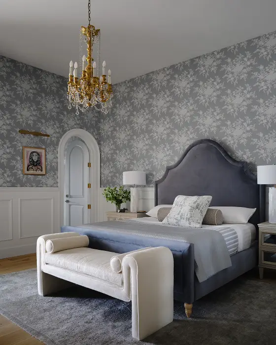 Elegant bedroom with oval door, tufted blue headboard, gold chandelier, and gray floral accent wall with bench.