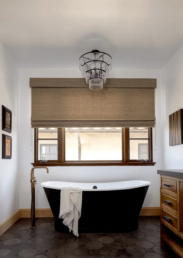 Elegant bathroom with black soaking tub, brass faucet, wood cabinetry, Roman shades, and hex tile flooring.