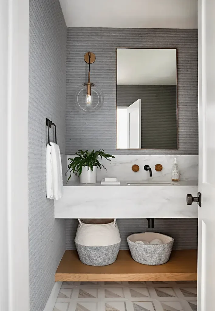 Modern powder room with marble vanity, gray textured wallpaper, brass fixtures, and woven baskets for storage.