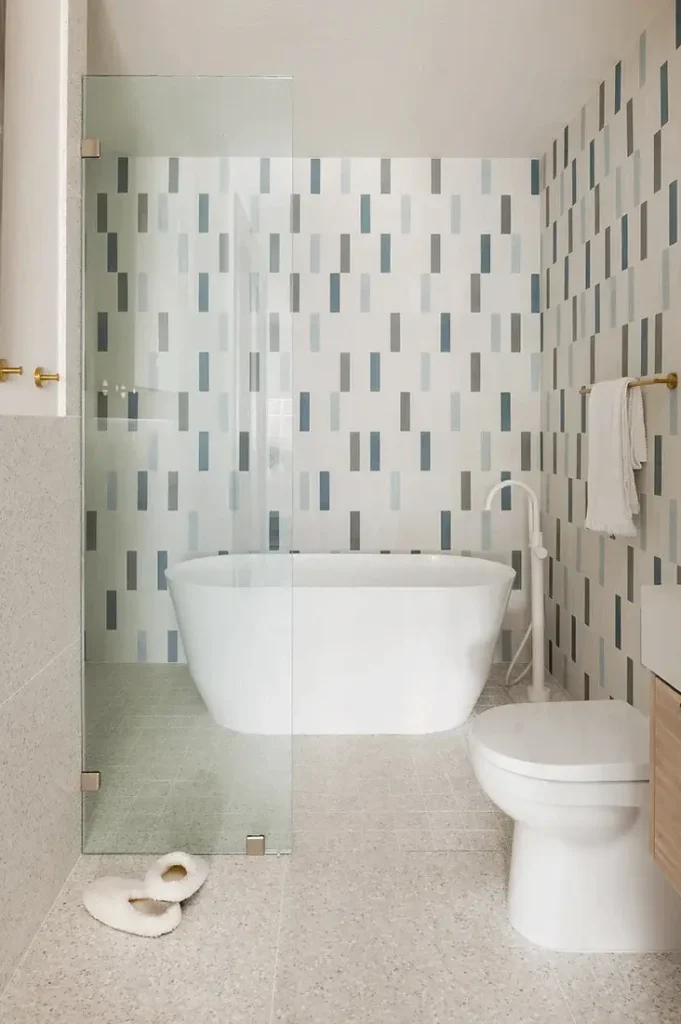 Modern bathroom with freestanding tub, terrazzo floor, blue tile accents, brass fixtures, and wood cabinetry.
