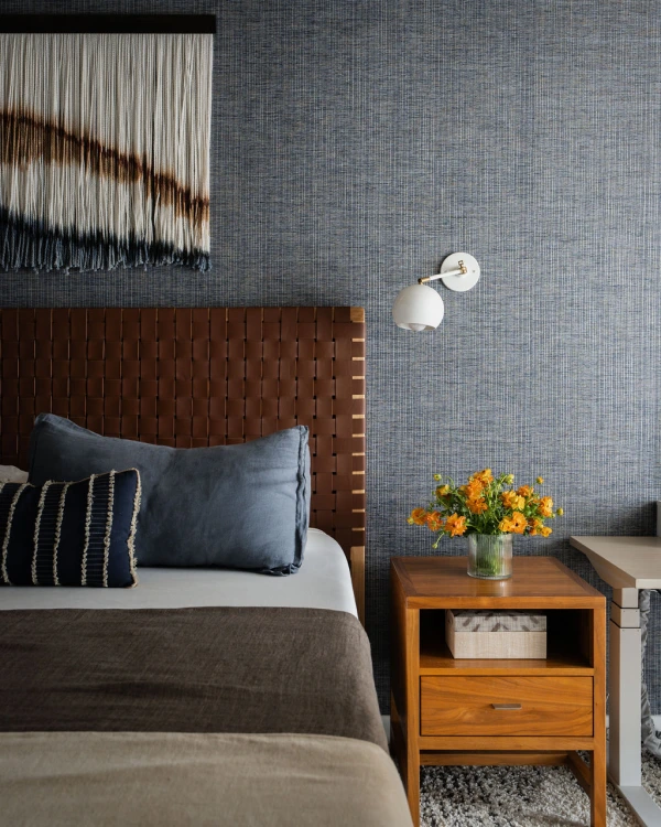 Bedroom corner with violet wallpaper, leather headboard, blue patterned pillows and wood nightstand with yellow flowers