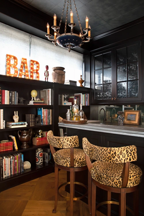 Home bar with black cabinetry, leopard print barstools, open shelves, chandelier, and illuminated marquee BAR letters.