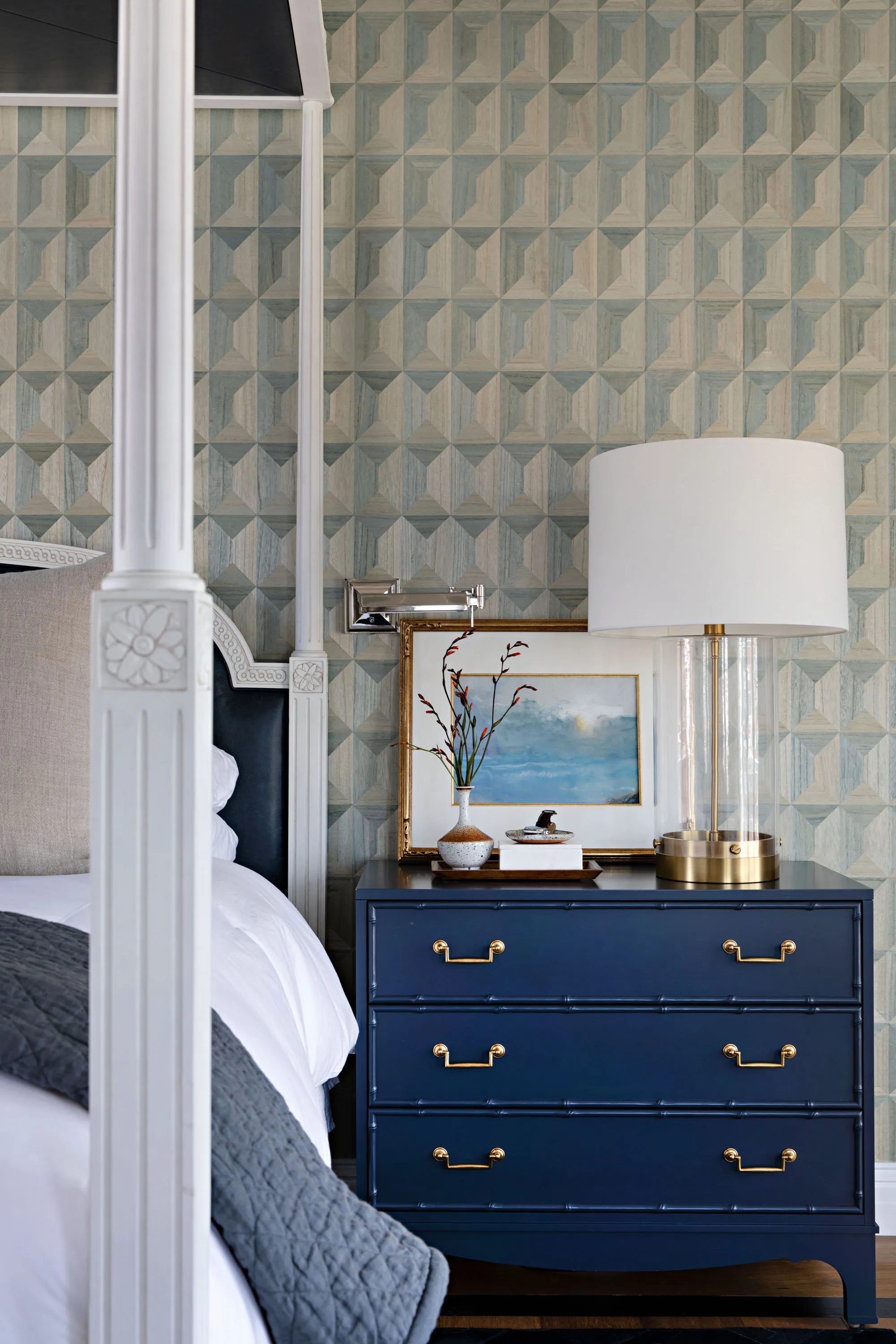 Bedside detail with a navy campaign chest, glass lamp and framed coastal painting