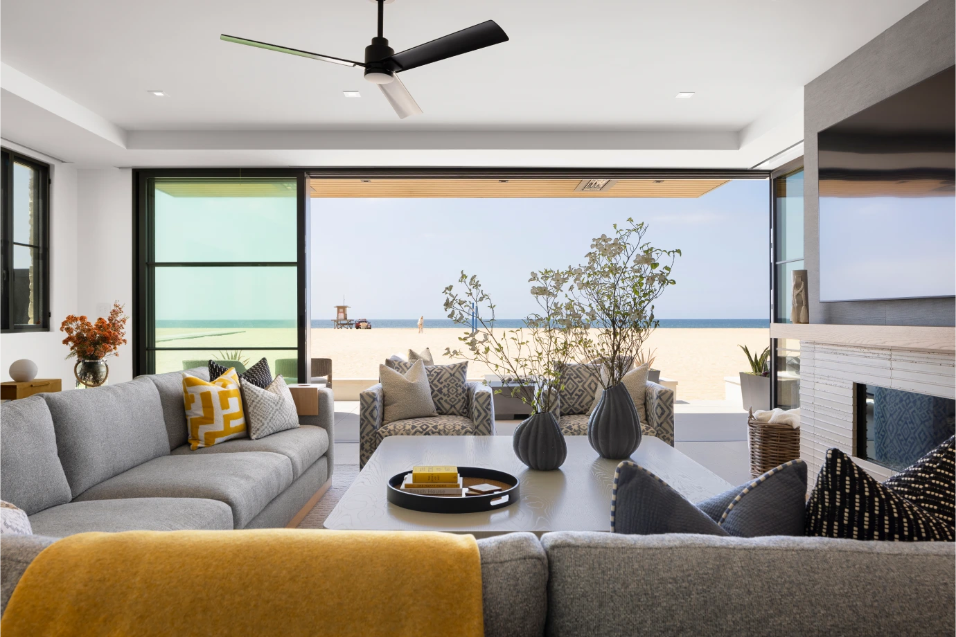 Ocean-front living room from Grace Blu's Interior Design Project The Happy Place with light gray couch with a yellow blanket and pillow and floor-to-ceiling glass doors that open directly onto the sand