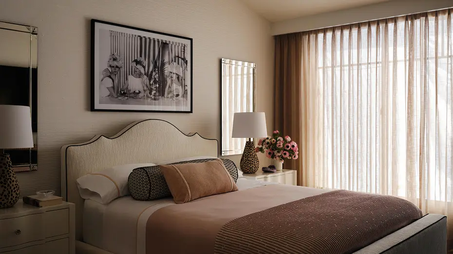 Bedroom featuring a queen-sized bed, photograph frame, and mirror. There are faded pink accents in the comforter and curtains with a beige wall