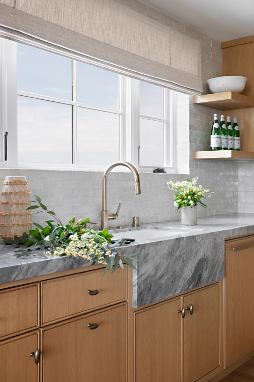 Kitchen from Grace Blu's Park Ave project with light gray marble counters, white tiling for the walls and light brown cabinets. There are windows placed just above the sink.