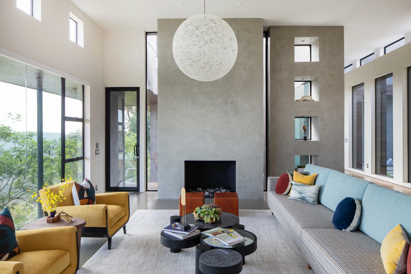Living room with glass wall, gray palette, colorful cushions, wood wall art, and a modern circular white pendant lamp.