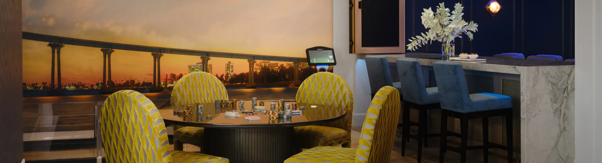 Chic game room with patterned yellow chairs, round poker table, and marble bar with blue stools.