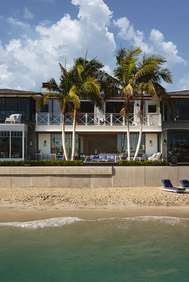 Modern beachfront home with palm trees and outdoor seating facing the ocean.