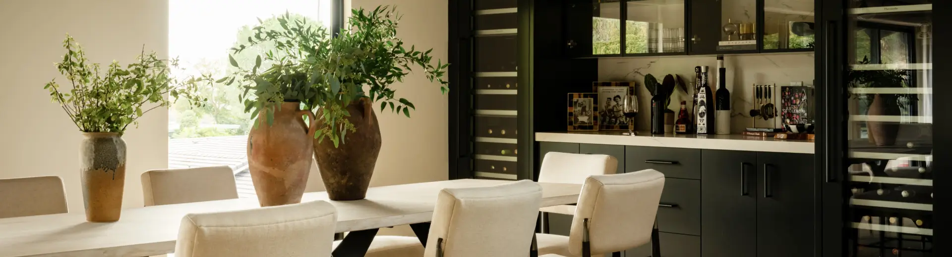 Modern dining space with large vases of greenery, beige chairs, and a sleek built-in bar and wine storage.