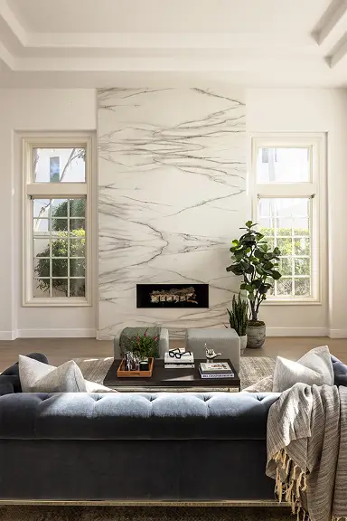 white living room with marble fireplace and gold accent above, flanked by symmetrical windows letting in natural light