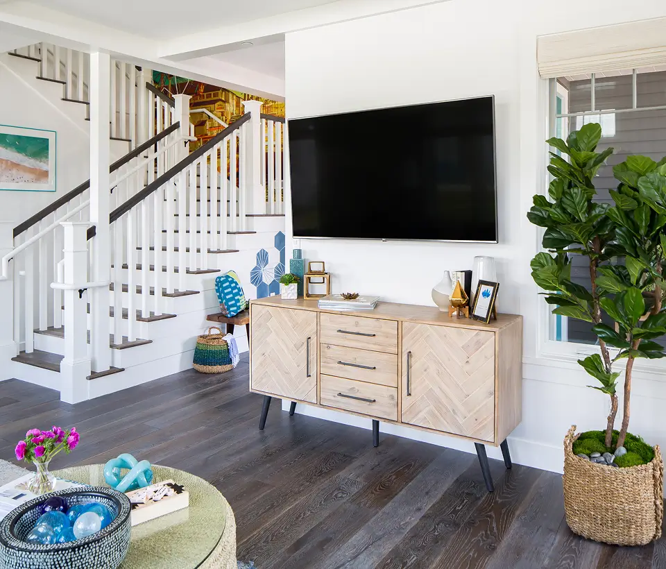 Bright living room with mounted TV, light wood console, indoor plant, and staircase with white railings and dark wood accents.