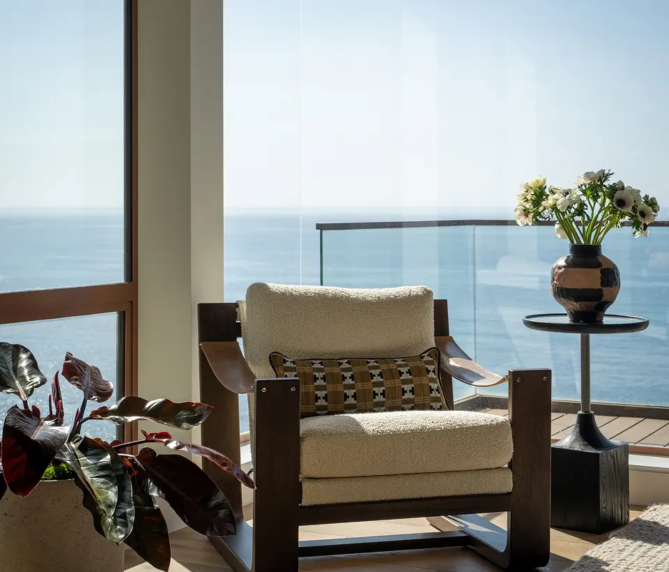 Cozy lounge chair with patterned cushion beside a vase of flowers, overlooking ocean views through glass balcony doors.