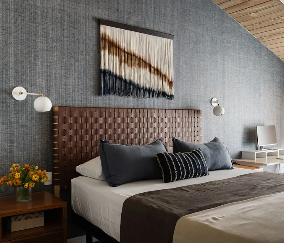 Modern bedroom with woven leather headboard, textured wall art, gray bedding, and wooden nightstand with fresh flowers.
