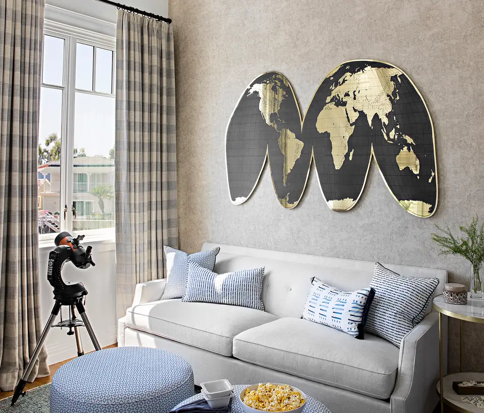 Cozy living room with gray sofa, blue patterned pillows, world map wall art, telescope by the window, and ottoman with popcorn.