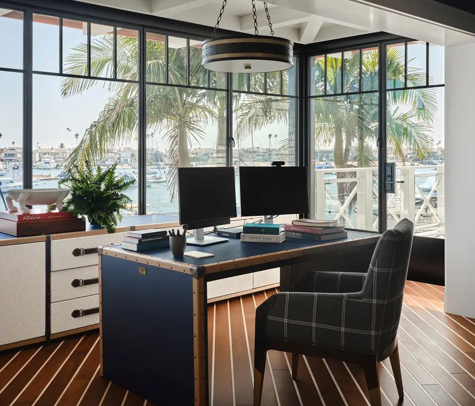 Stylish home office with waterfront view, dual monitors, plaid chair, wood flooring, and large windows overlooking boats and palm trees.