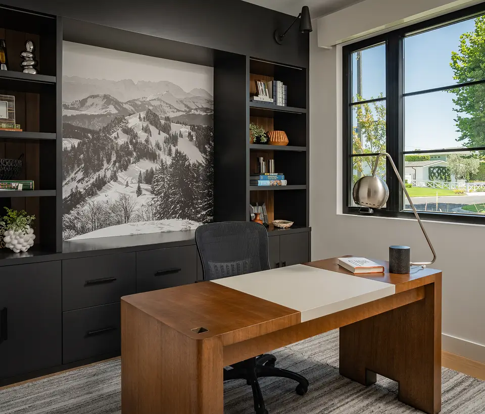 Modern home office with wooden desk, black shelving, mountain landscape wall art, and large window bringing in natural light.