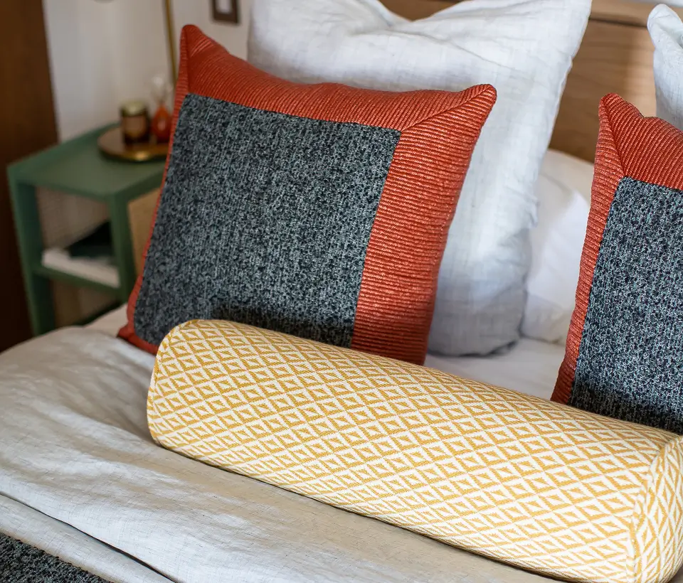 Close-up of bed with textured pillows in gray and rust tones, paired with a yellow patterned bolster cushion.