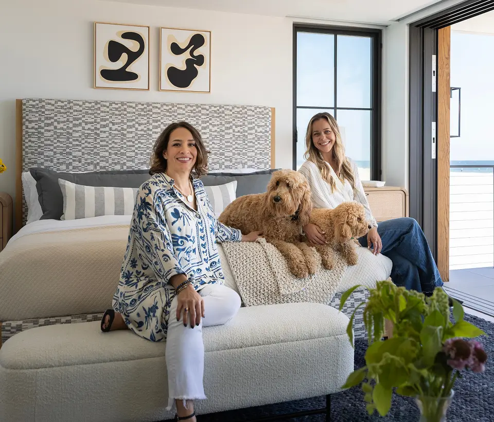 Rona, founder of Grace Blu, and designer Courtney sit on a bed with two dogs in a modern bedroom with patterned headboard and wall art.