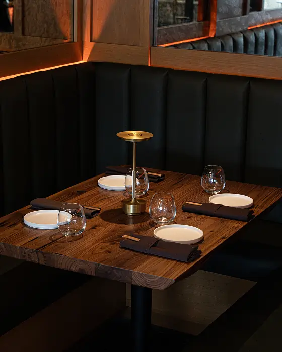 Rustic wooden restaurant table set for four with white plates, glassware, brown napkins, and a gold table lamp.
