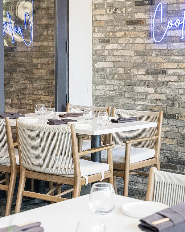 Light-filled table for four with rope-back wood chairs, white tabletop, and brick wall with neon glow at Yuzu Sushi.