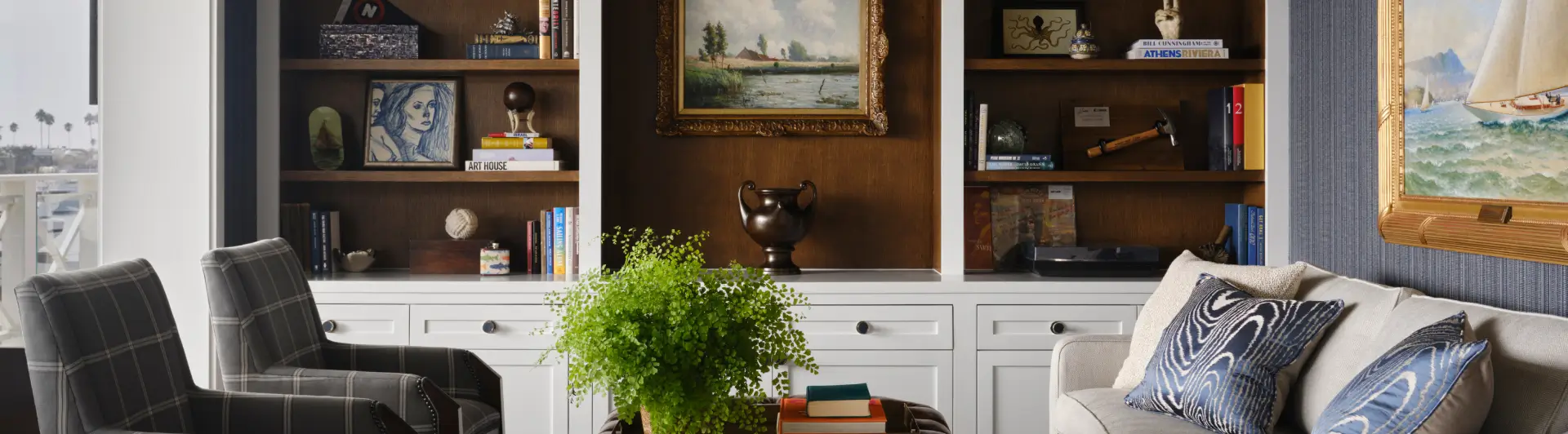 Cozy living room with bookshelves, framed art, leather ottoman, and a green plant centerpiece.