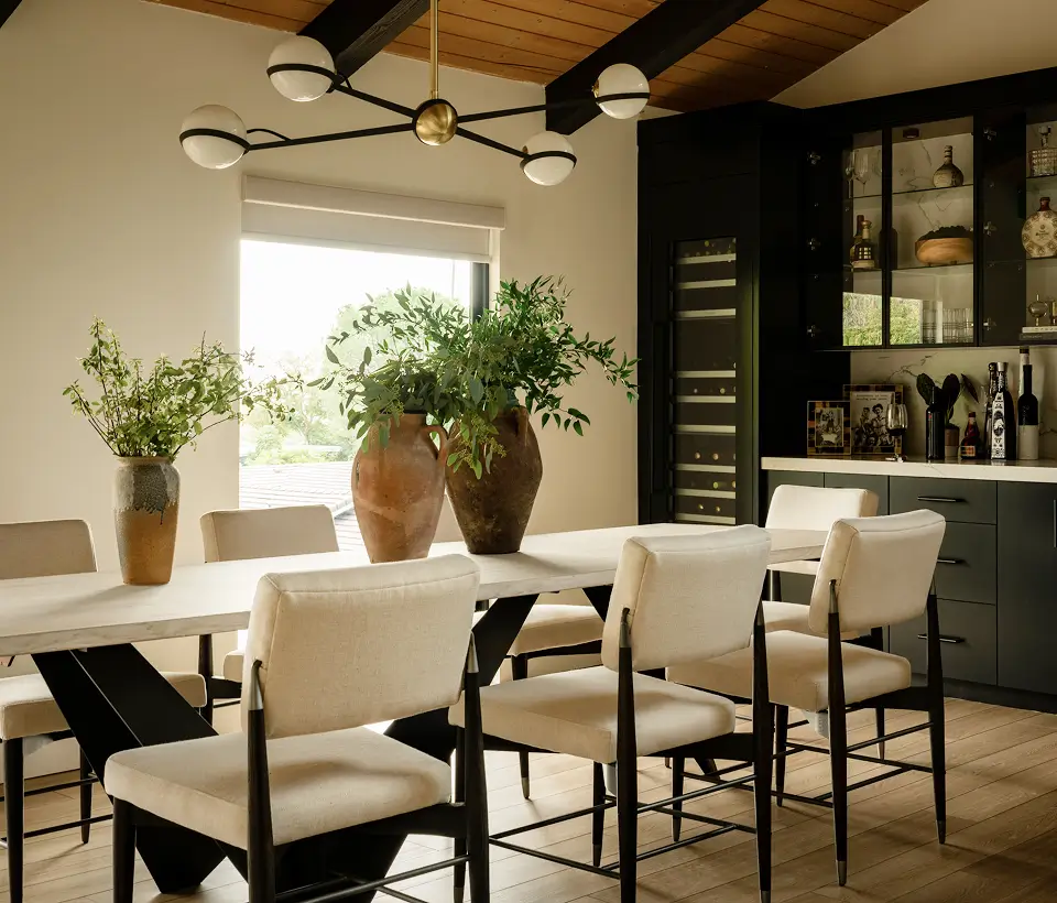 Modern dining room with beige chairs, greenery in vases, black cabinetry, and a sleek wine storage wall.