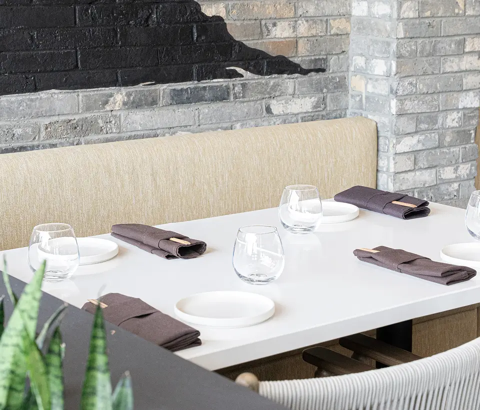 Bright restaurant table with beige booth seating, white plates, glassware, dark napkins, and exposed brick wall backdrop.