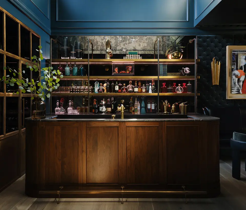 Elegant home bar with dark wood cabinetry, mirrored shelves stocked with liquor bottles, gold accents, and moody blue walls.